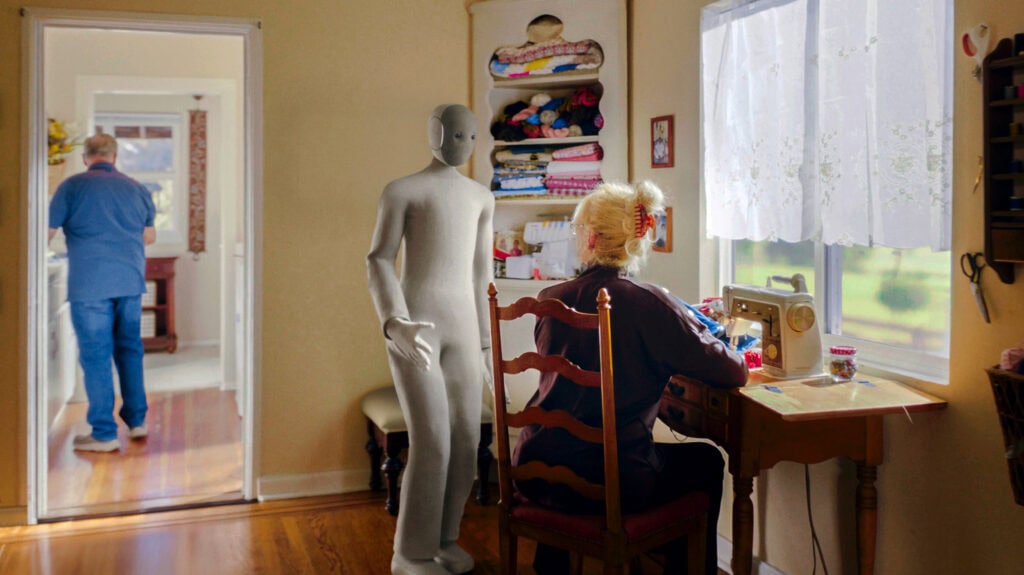 Foto van NEO die in een huiskamer staat naast een zittende mevrouw met grijs haar.